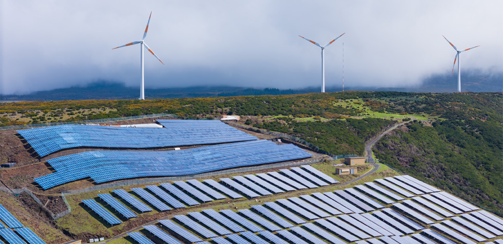 Photograph of a solar panels on a hill with wind turbines behind